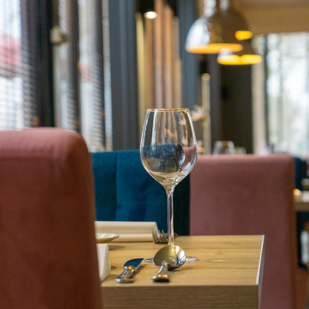Gedekte restauranttafel met wijnglas en bestek, omringd door roodbruine en blauwe stoelen.
