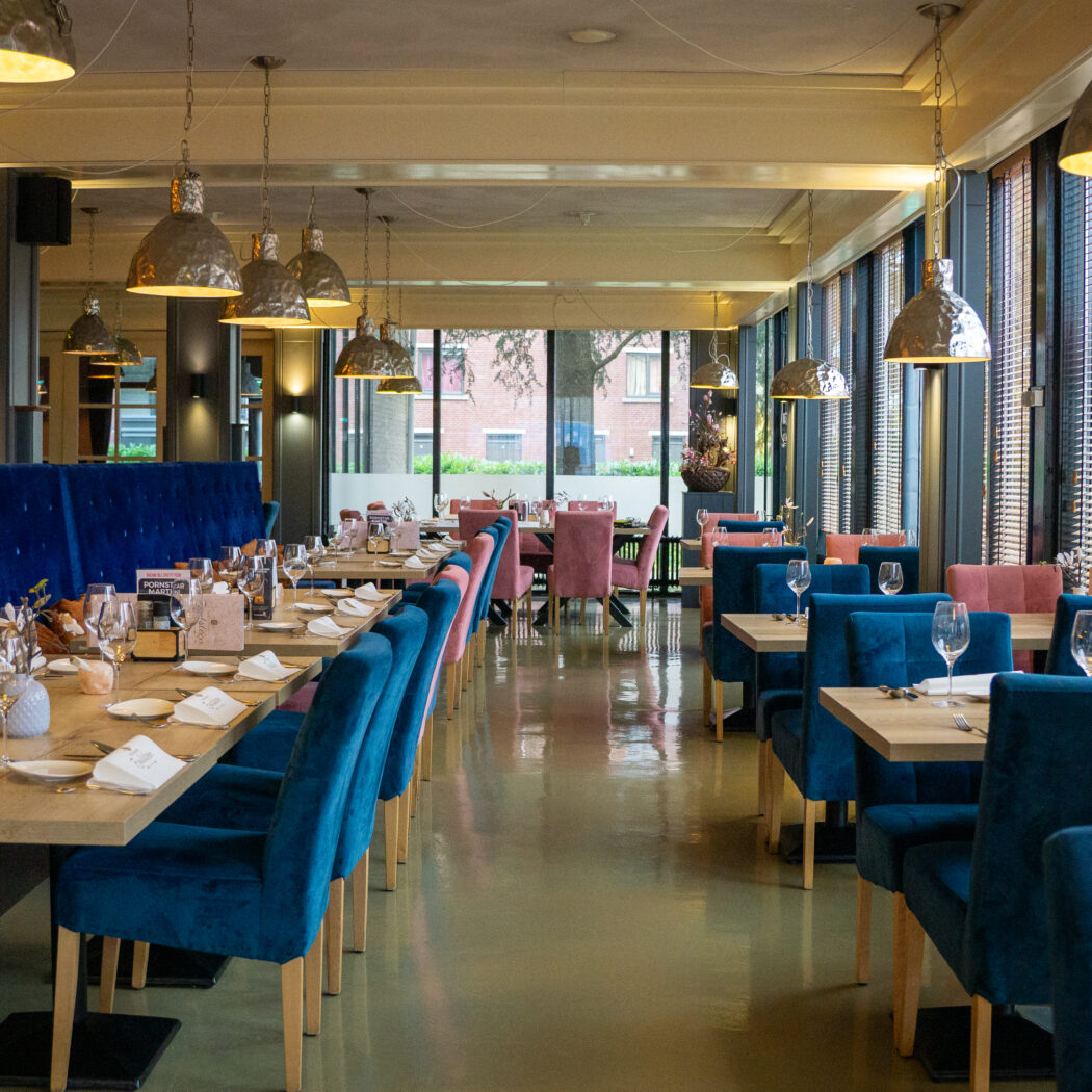 Sfeervol restaurant met blauwe fluwelen banken, blauwe en roze stoelen, houten tafels en moderne verlichting.
