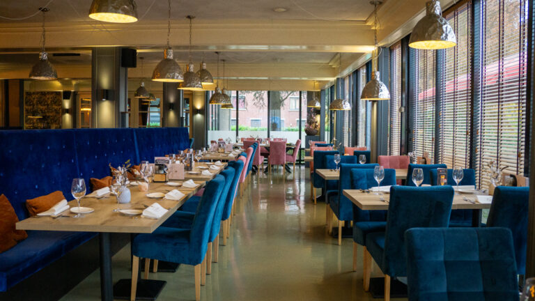 Sfeervol restaurant met blauwe fluwelen banken, blauwe en roze stoelen, houten tafels en moderne verlichting.