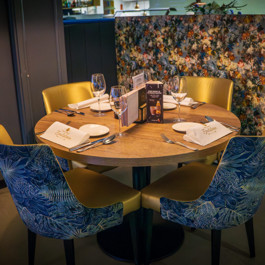 Ronde houten restauranttafel gedekt voor vier met blauw-goudgele stoelen en Zuidlanden servetten, tegen bloemrijk behang.