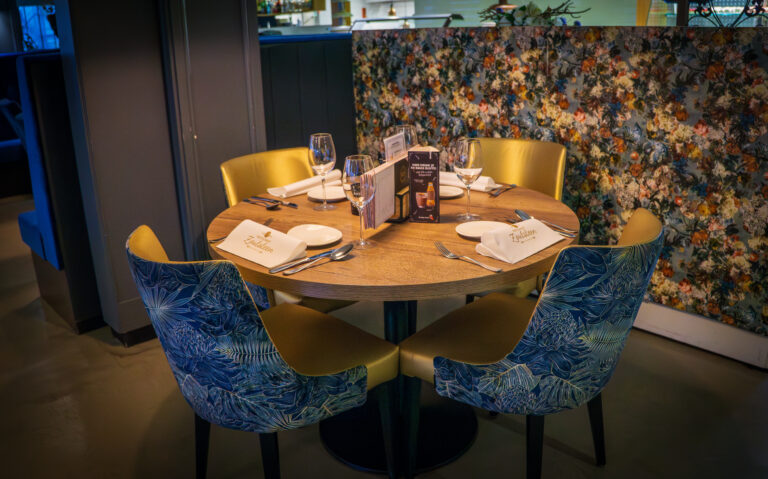 Ronde houten restauranttafel gedekt voor vier met blauw-goudgele stoelen en Zuidlanden servetten, tegen bloemrijk behang.