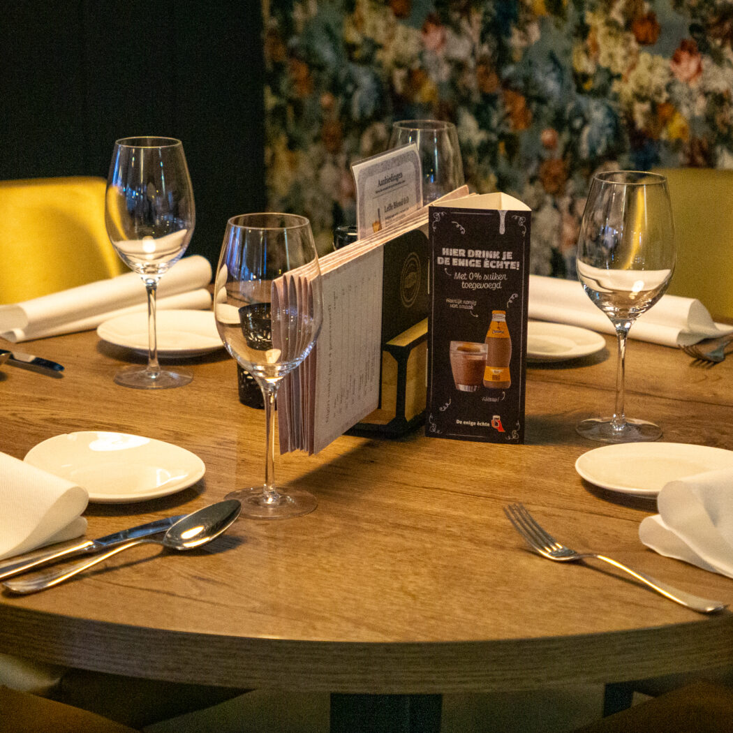 Ronde houten restauranttafel gedekt met bestek, glazen, borden, Restaurant Zoutsteen servetten en menukaart, bij gouden stoelen.