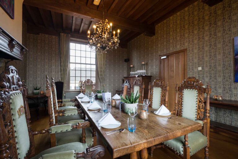 Lange houten eetkamertafel gedekt voor diner, omringd door groene antieke stoelen onder een kroonluchter.