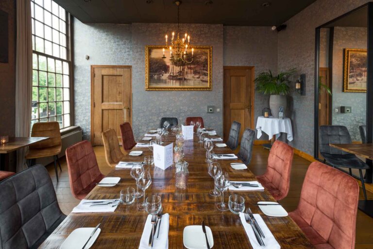 Lange rustieke houten eettafel gedekt met bestek en glazen, omringd door diverse stoelen in een elegant restaurant.
