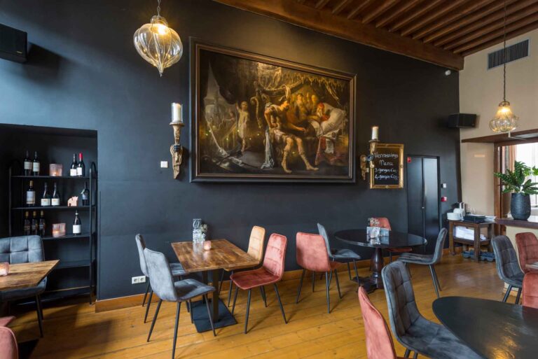 Sfeervol restaurantinterieur met donkergrijze muren, groot schilderij, houten vloer, kleurrijke stoelen en Verrassings Menu bord.