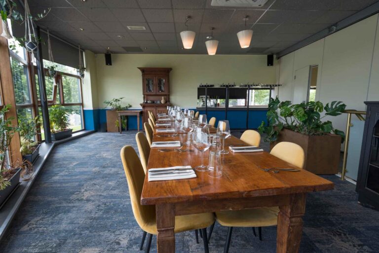 Gedekte lange, rustieke houten eettafel met gele stoelen in een lichte zaal met grote ramen en planten.