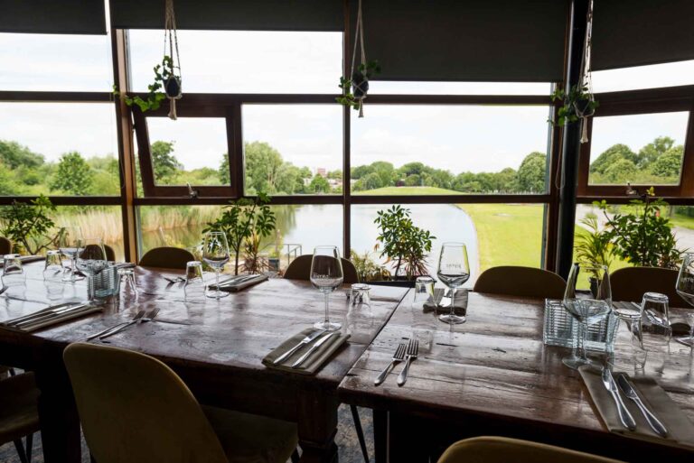 Gedekte houten tafels met wijnglazen en bestek in een restaurant met panoramisch uitzicht op een meer en groen landschap.