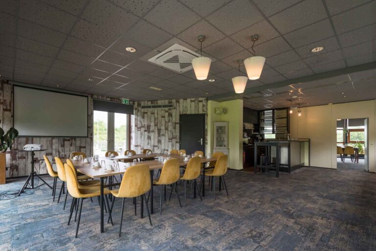 Lichtrijke vergaderzaal met lange houten tafel, mosterdgele stoelen, projectiescherm en een bar.