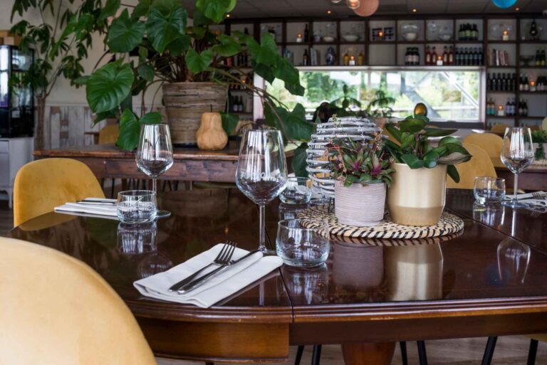 Gedekte houten eettafel met gele stoelen, glazen, bestek en potplanten in een sfeervol restaurantinterieur.