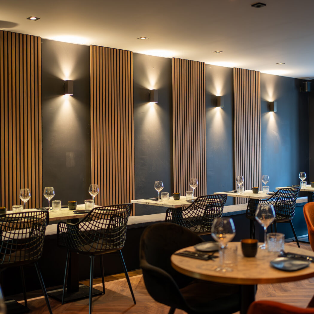 Modern restaurantinterieur met houten wandpanelen, sfeervolle verlichting en gedekte tafels met diverse stoelen.