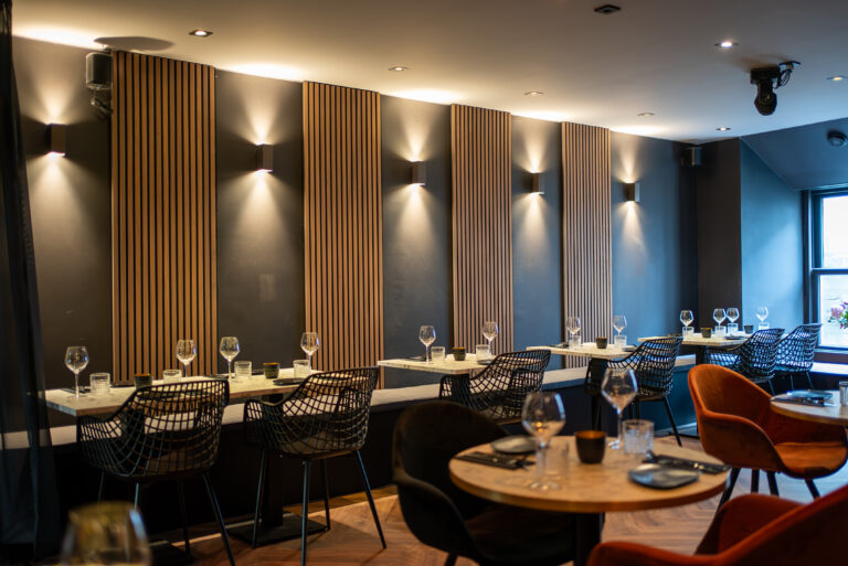 Modern restaurantinterieur met houten wandpanelen, sfeervolle verlichting en gedekte tafels met diverse stoelen.