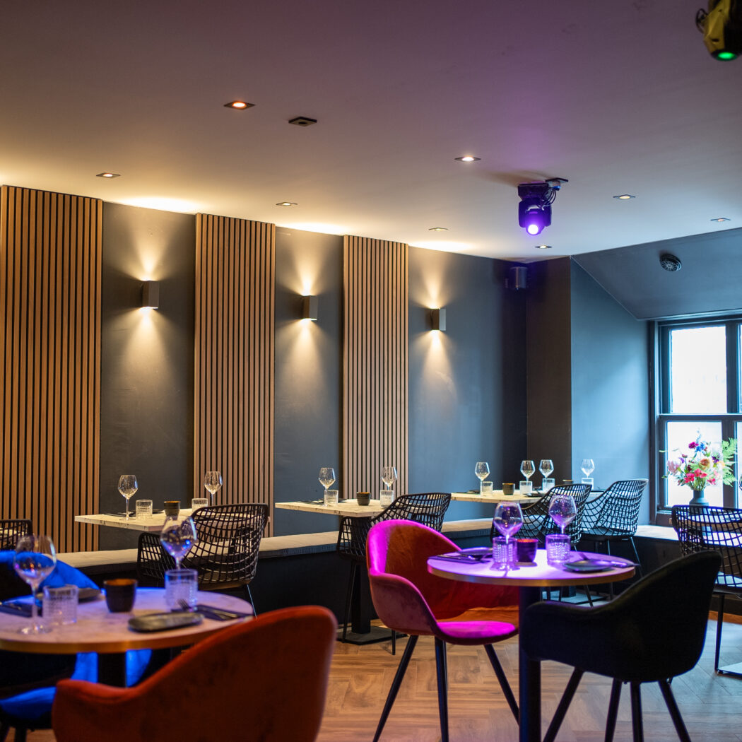 Sfeervol restaurant met gedekte tafels, zwarte muren, houten accenten en kleurrijke stoelen.