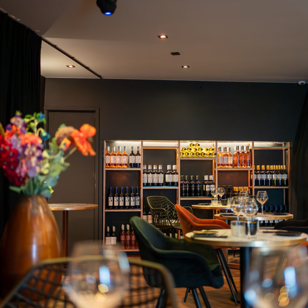Sfeervol restaurantinterieur met verlichte wijnrekken, ronde tafels met velvet stoelen en een vaas met bloemen.