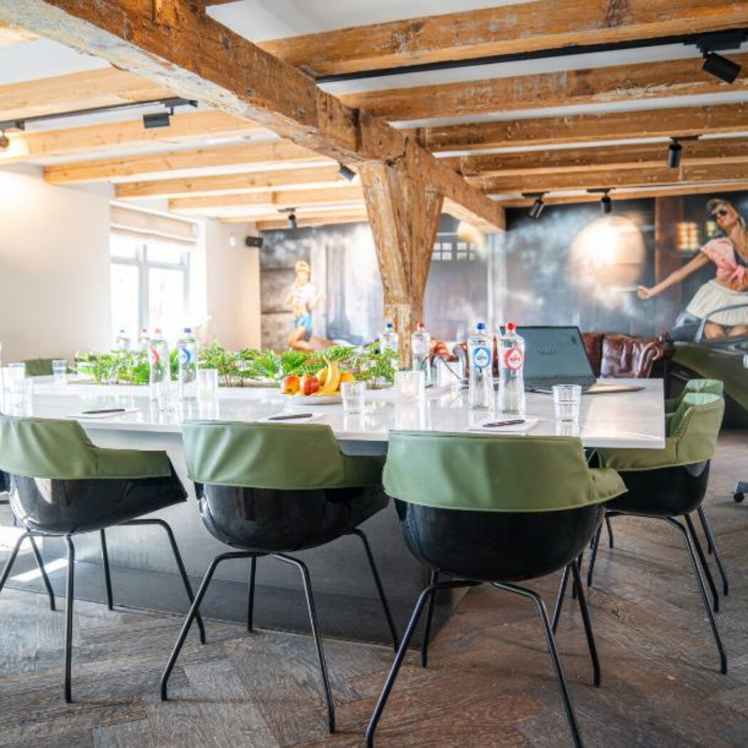 Lichte vergaderzaal met lange witte tafel, zwarte stoelen met groene rugleuningen, houten balken en levendige muurschilderingen.