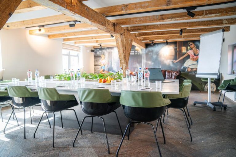 Lichte vergaderzaal met lange witte tafel, zwarte stoelen met groene rugleuningen, houten balken en levendige muurschilderingen.