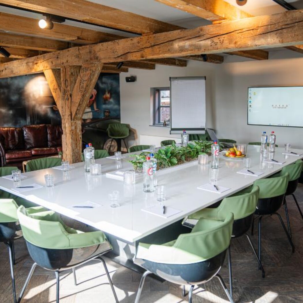 Sfeervolle vergaderzaal met een lange witte tafel, frisgroene stoelen, houten balken, scherm en lederen banken.