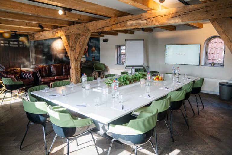 Sfeervolle vergaderzaal met een lange witte tafel, frisgroene stoelen, houten balken, scherm en lederen banken.