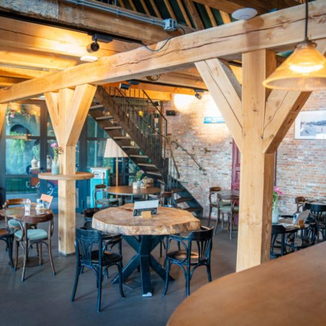 Gezellig restaurantinterieur met robuuste houten balken, bakstenen muren, diverse tafels, een koperen haard en trap.