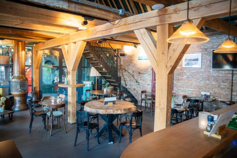 Gezellig restaurantinterieur met robuuste houten balken, bakstenen muren, diverse tafels, een koperen haard en trap.