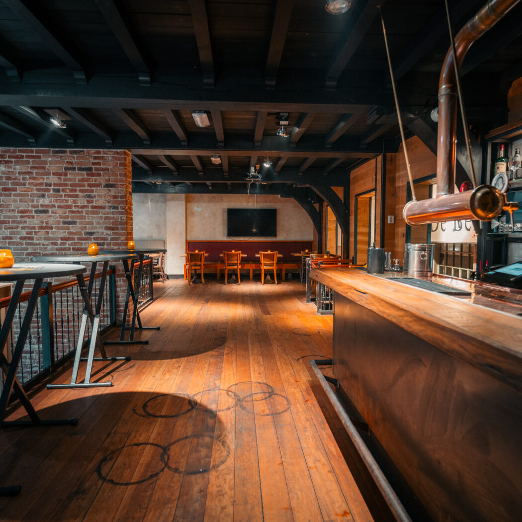 Rustiek interieur van een lege bar met houten vloer, bakstenen muur, hoge tafels en koperen tap.