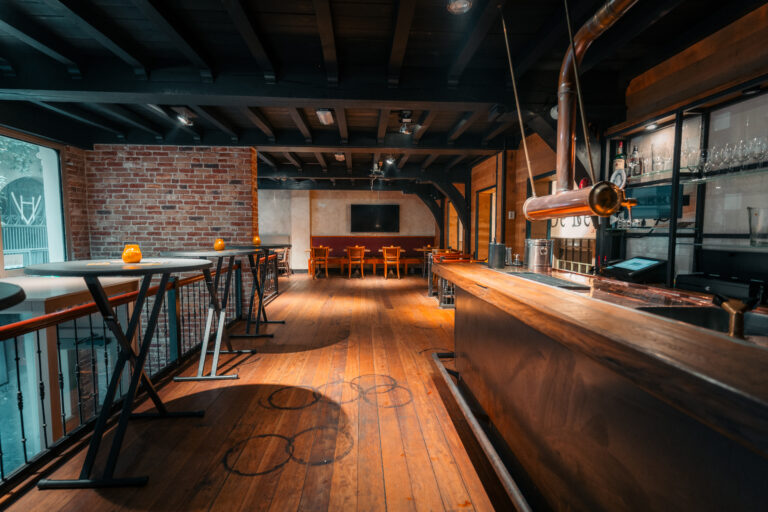 Rustiek interieur van een lege bar met houten vloer, bakstenen muur, hoge tafels en koperen tap.