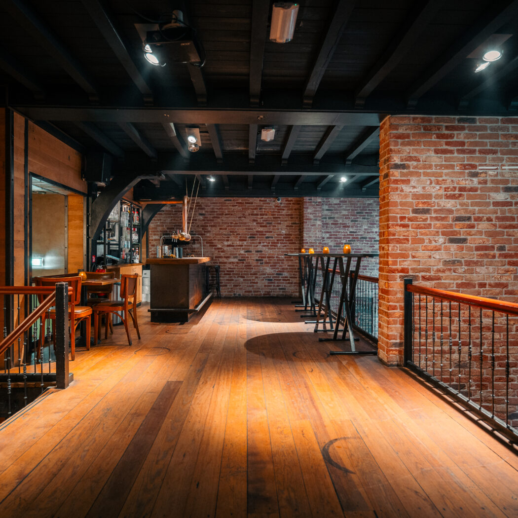 Ruime evenementenlocatie met houten vloer, bakstenen muren, een bar en statafels met kaarslicht.