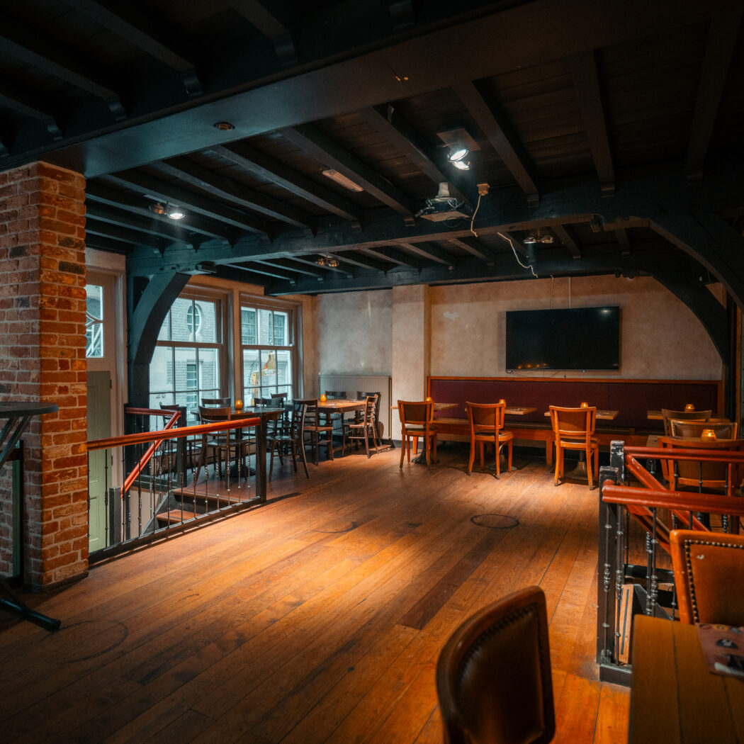 Sfeervol interieur van bar/restaurant met houten vloer, bakstenen muur, balkenplafond en diverse zitjes.
