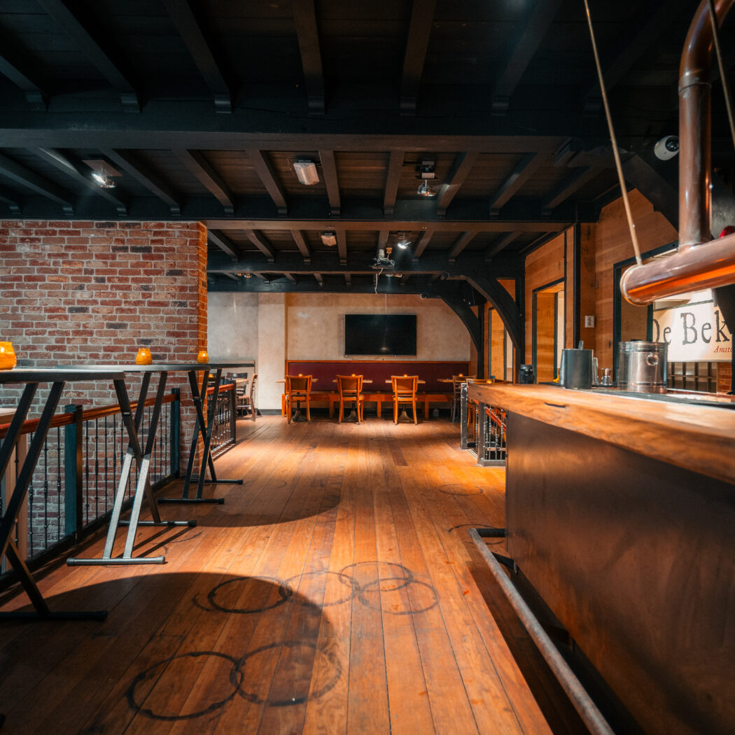 Warm interieur van café De Bek met lange houten bar, koperen biertap, hoge tafels en zithoek.