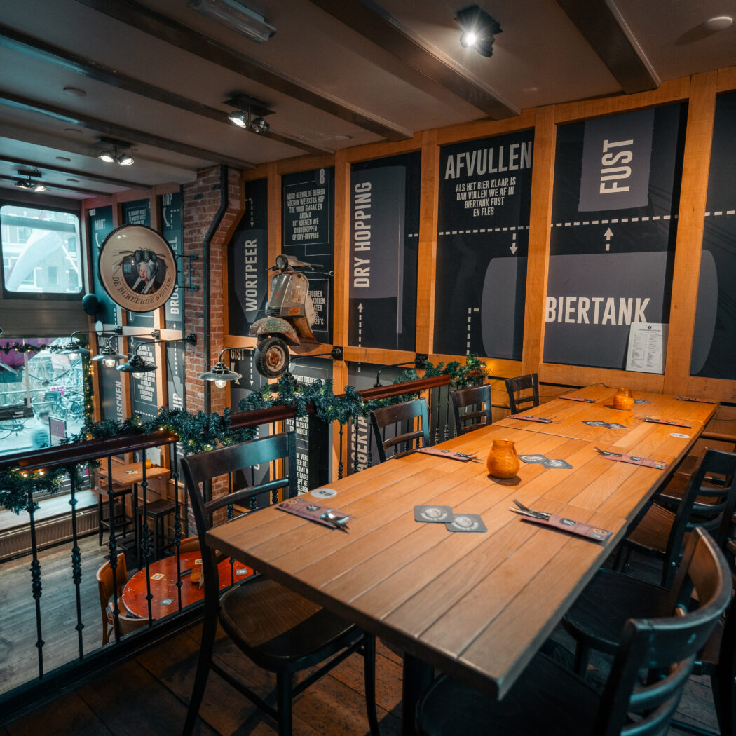 Sfeervol interieur van een brouwerij-pub met lange houten tafel, scooter aan de wand, en muurpanelen over BIERTANK.