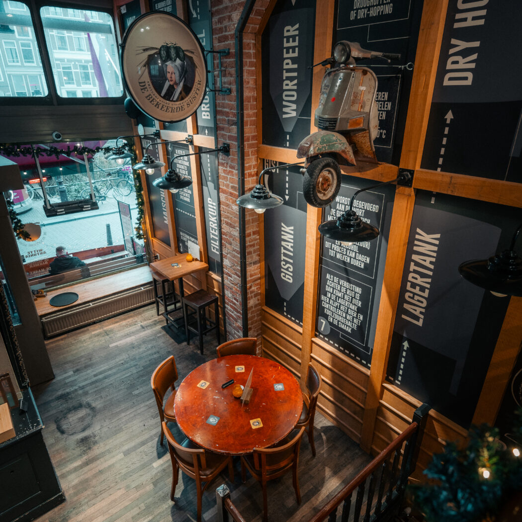 Sfeervol brouwerijcafé interieur met houten tafels, een vintage Vespa en wandpanelen die het bierbrouwproces uitleggen, met DE BEKEERDE SUSTER.