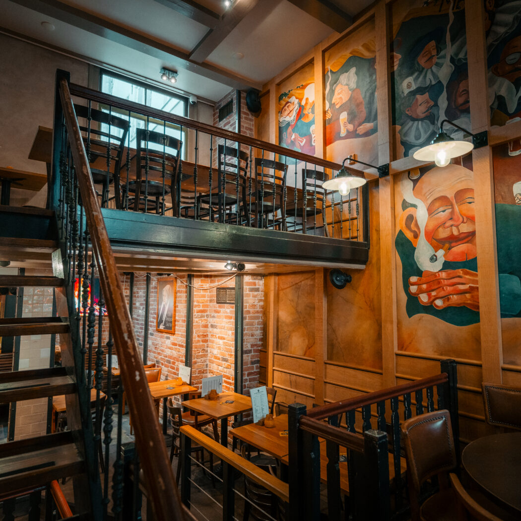 Interieur van een pub of restaurant met houten trap naar een mezzanine vol tafels en stoelen, en muren met kleurrijke muurschilderingen.