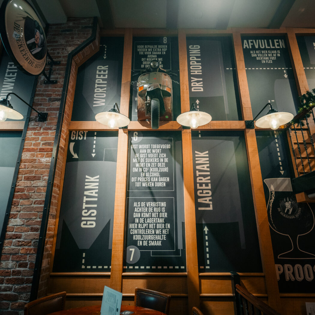 Muur in een brouwerij met infographic van het bierbrouwproces (Gisttank, Lagertank), een gemonteerde scooter en lampen.