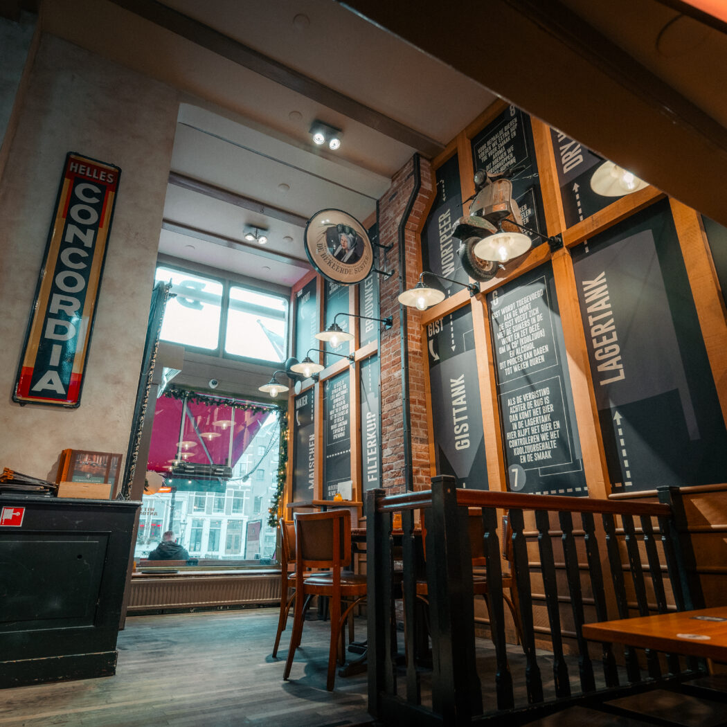Gezellig interieur van een brouwerijproeflokaal met houten tafels, bierbrouw-infopanelen en een HELLES CONCORDIA wandbord.