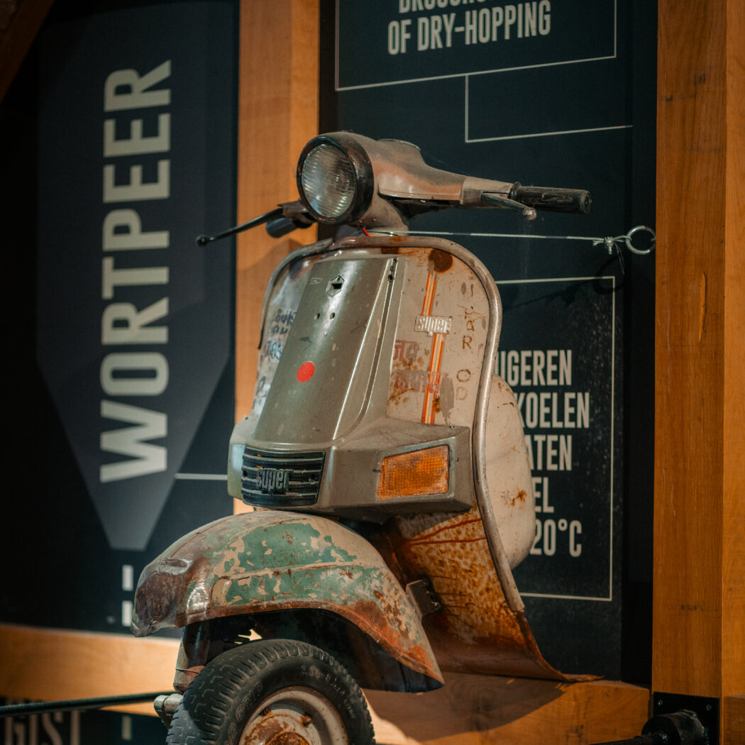 Roestige Super scooter met groen spatbord, als decoratie hangend tegen een donkere wand met brouwerijteksten.