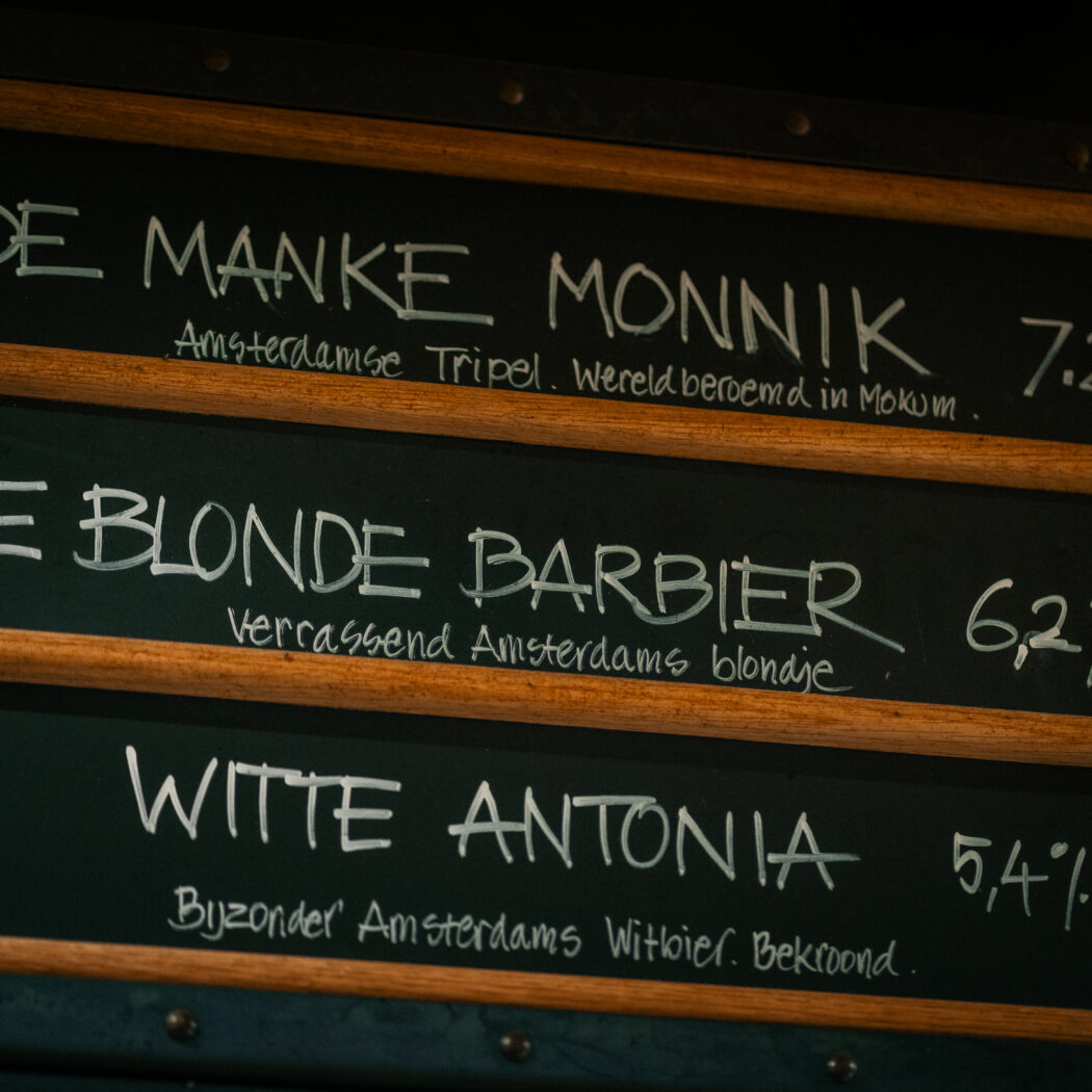 Zwart krijtbord menu met Amsterdamse bieren: De Manke Monnik (7.2%, 6,90), De Blonde Barbier (6.2%, 6,90) en Witte Antonia (5.4%, 6,90).