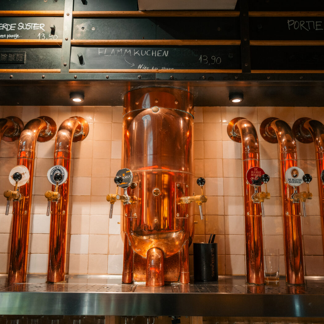 Glimmende koperen biertaps en een brouwvat in een bar, met krijtborden die Proeverij De Bekeerde Suster tonen.