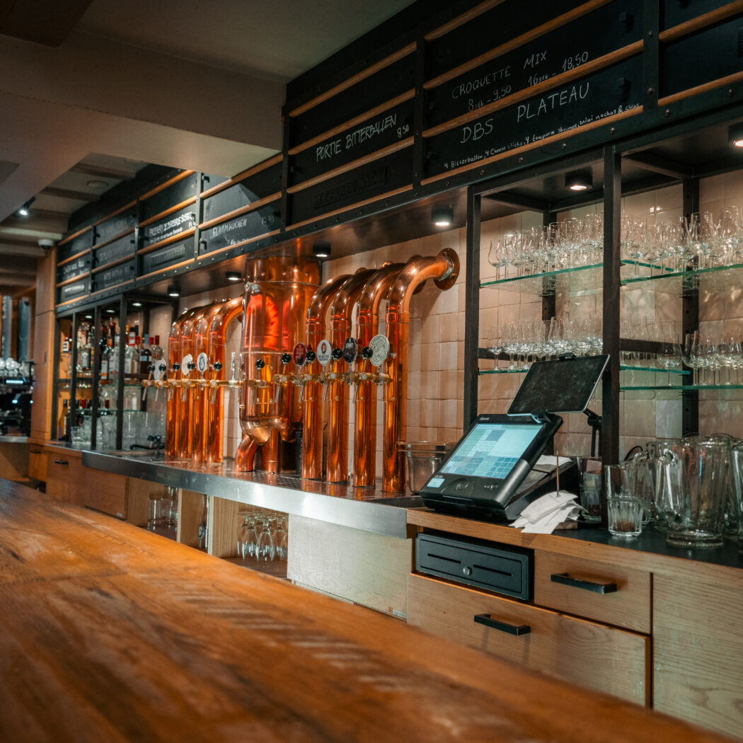 Koperen biertaps en een modern kassasysteem op een lange houten bar, met CROQUETTE MIX op het menu.