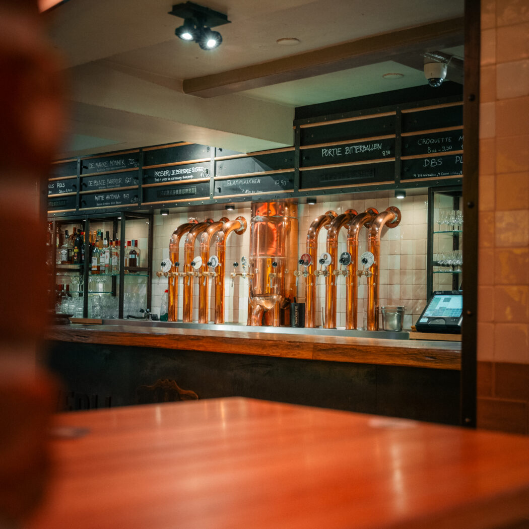 Interieur van een bar met rijen koperen biertaps, een menubord met Portie Bitterballen en gedistilleerd achter de toonbank.