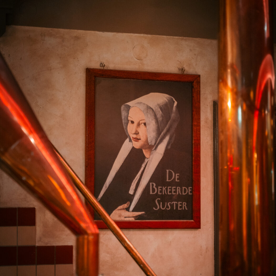 Ingelijst portret van een vrouw met wit kapje en de tekst De Bekeerde Suster, geflankeerd door glimmende koperen pijpen.
