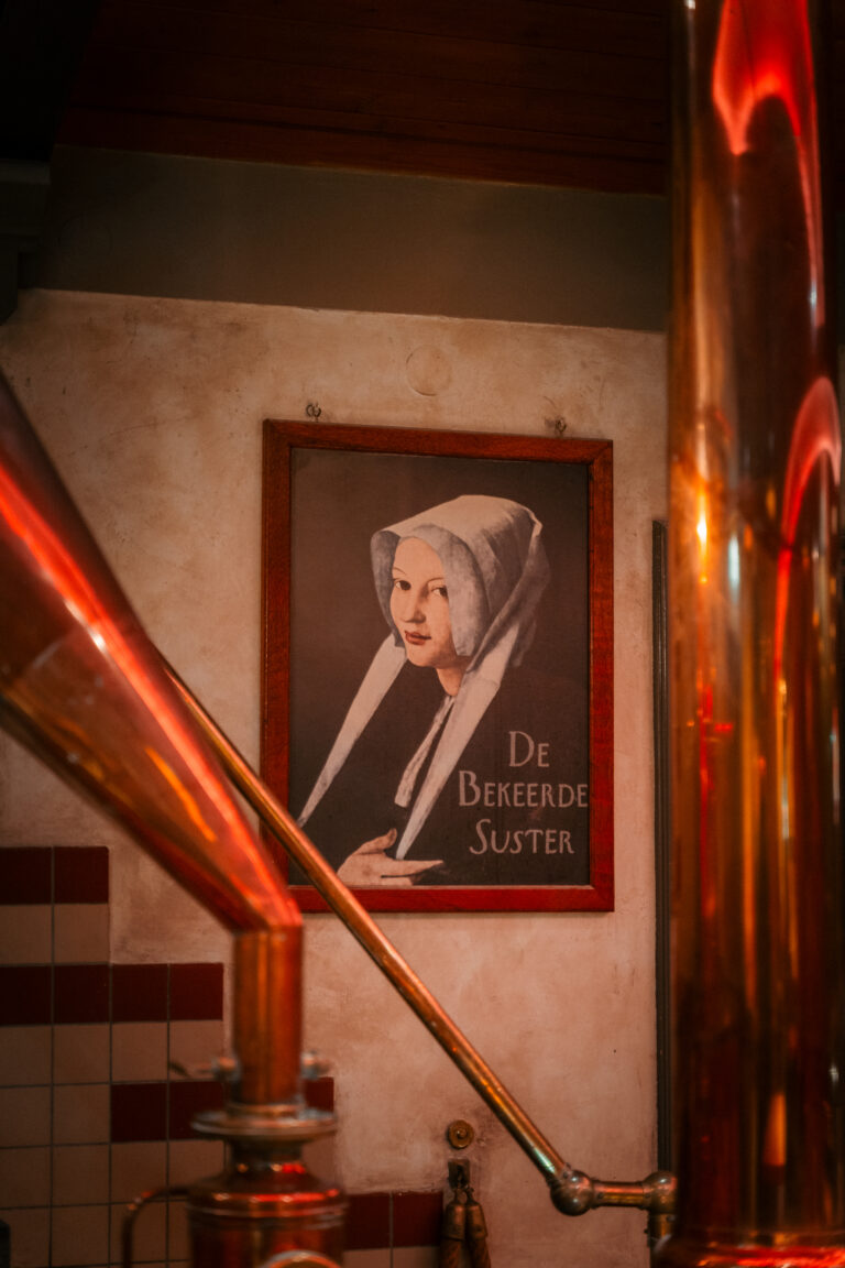 Ingelijst portret van een vrouw met wit kapje en de tekst De Bekeerde Suster, geflankeerd door glimmende koperen pijpen.