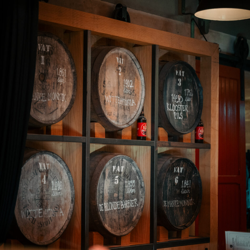 Zes grote houten vaten, elk met een nummer en naam zoals KLOOSTER PILS, op een houten rek met twee rode flessen.