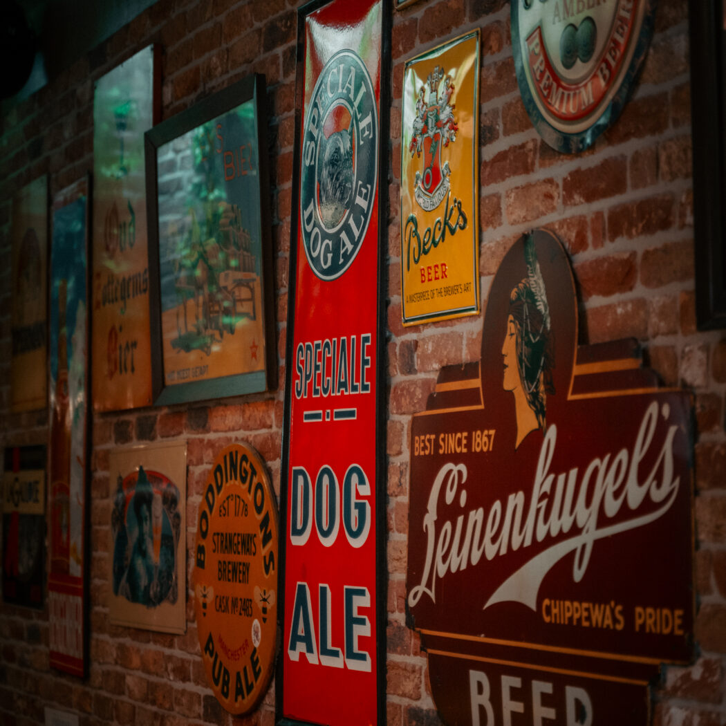Bakstenen muur met een verzameling vintage bierborden, waaronder Speciale Dog Ale, Leinenkugels en Becks.