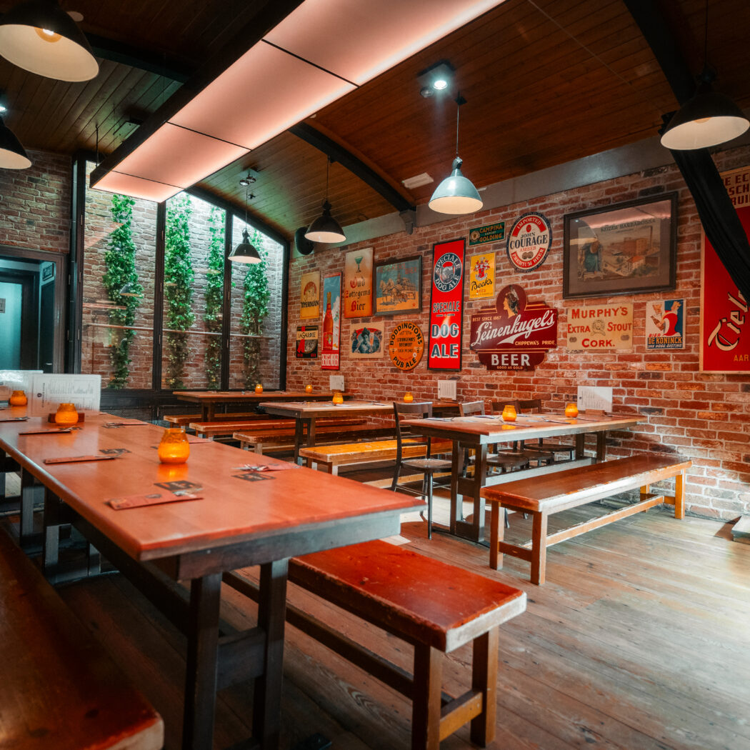 Gezellig café-interieur met houten tafels en banken, bakstenen muren vol vintage bierposters en oranje kaarslicht.