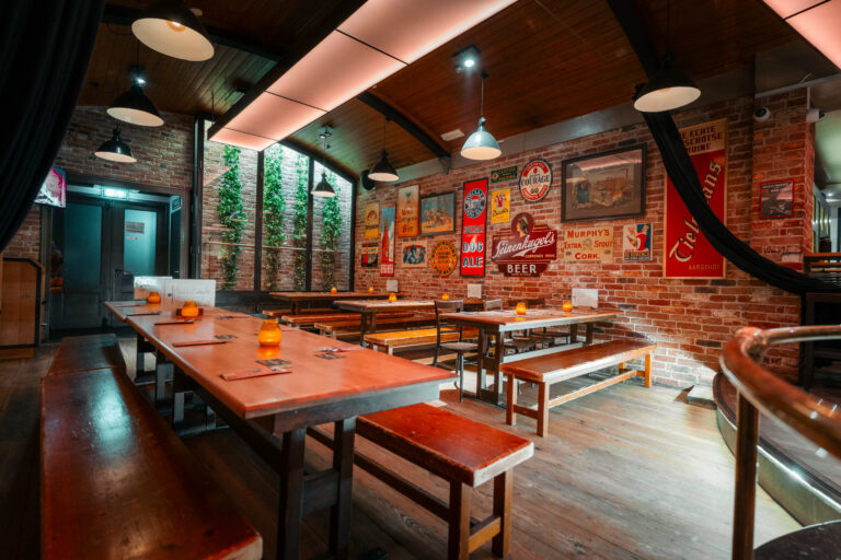 Gezellig café-interieur met houten tafels en banken, bakstenen muren vol vintage bierposters en oranje kaarslicht.