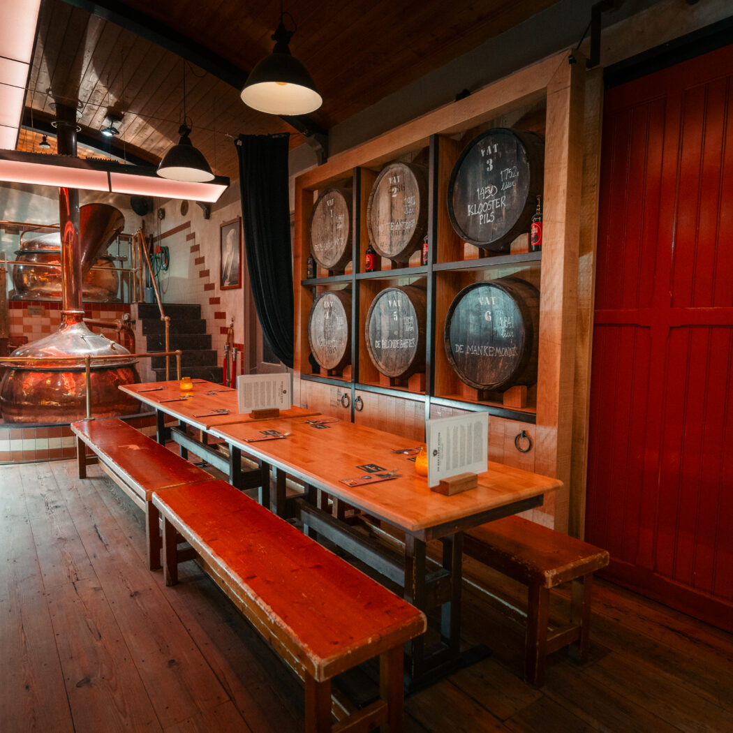 Sfeervolle brouwerij met glimmende koperen ketels, lange houten tafels, wand met vaten waaronder Klooster Pils, en een grote rode schuifdeur.