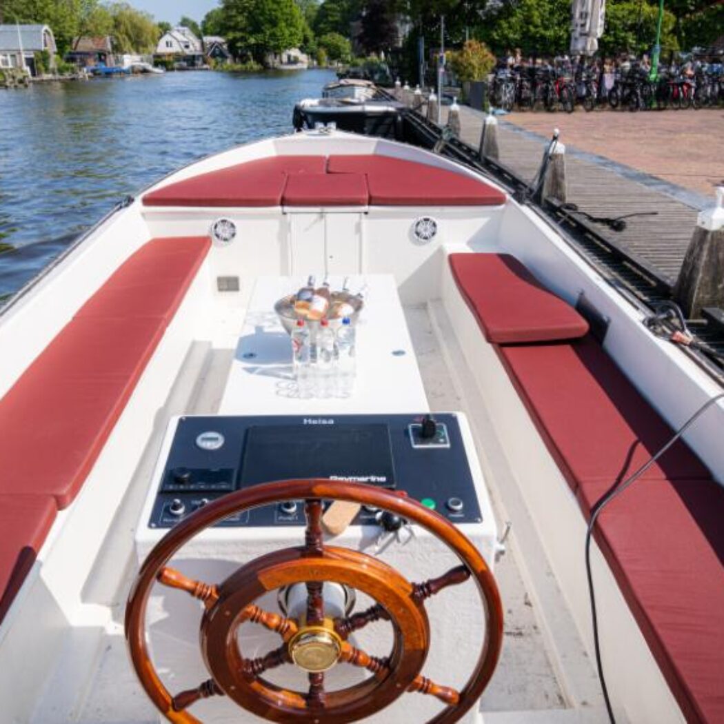 Witte sloep met roodbruine kussens, houten stuurwiel en bedieningspaneel, afgemeerd aan een houten steiger met drankjes op tafel.