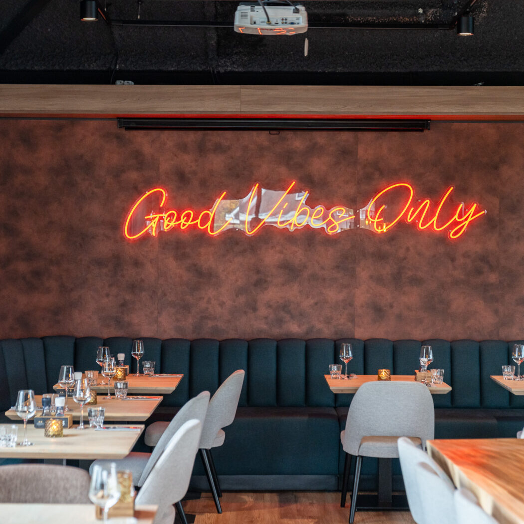 Sfeervol restaurantinterieur met een oranje neon Good Vibes Only bord, donkerblauwe banken en houten tafels.