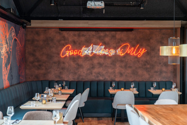 Sfeervol restaurantinterieur met een oranje neon Good Vibes Only bord, donkerblauwe banken en houten tafels.