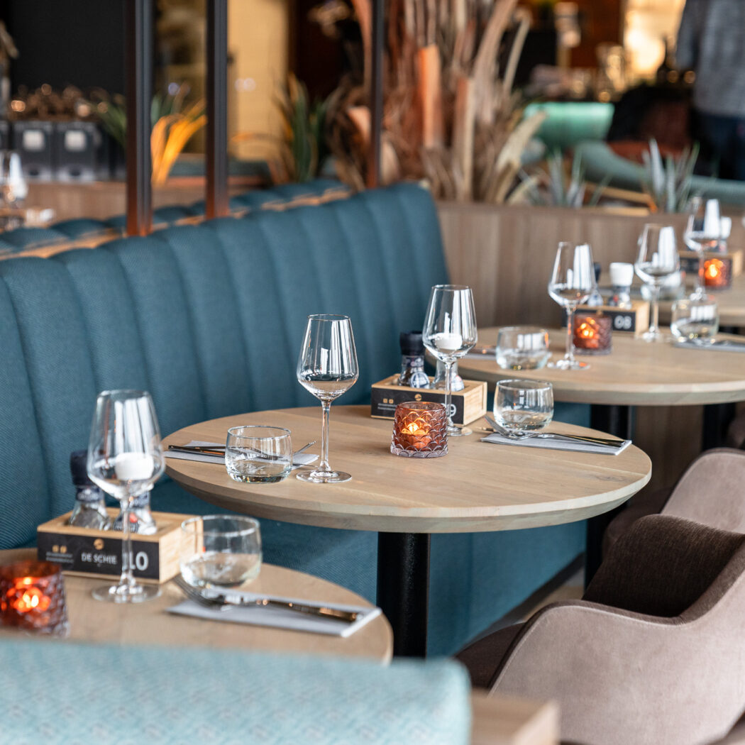 Gedekte tafels met wijnglazen, waterglazen en brandende kaarsen in een modern restaurant met teal banken.