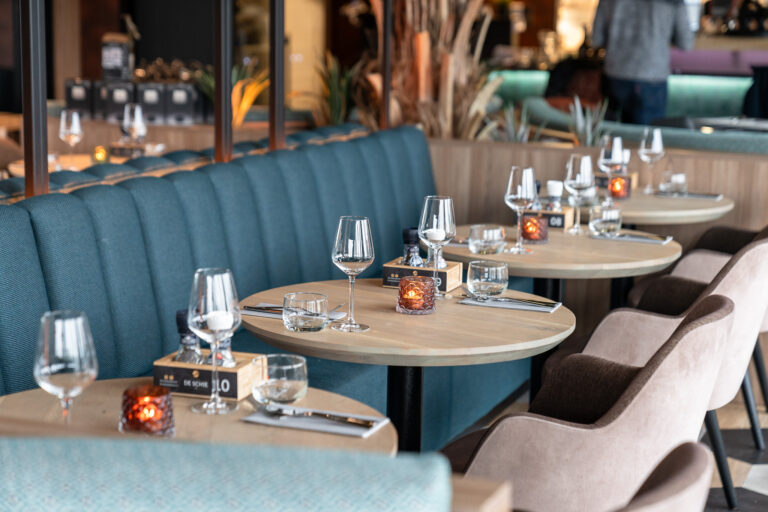 Gedekte tafels met wijnglazen, waterglazen en brandende kaarsen in een modern restaurant met teal banken.